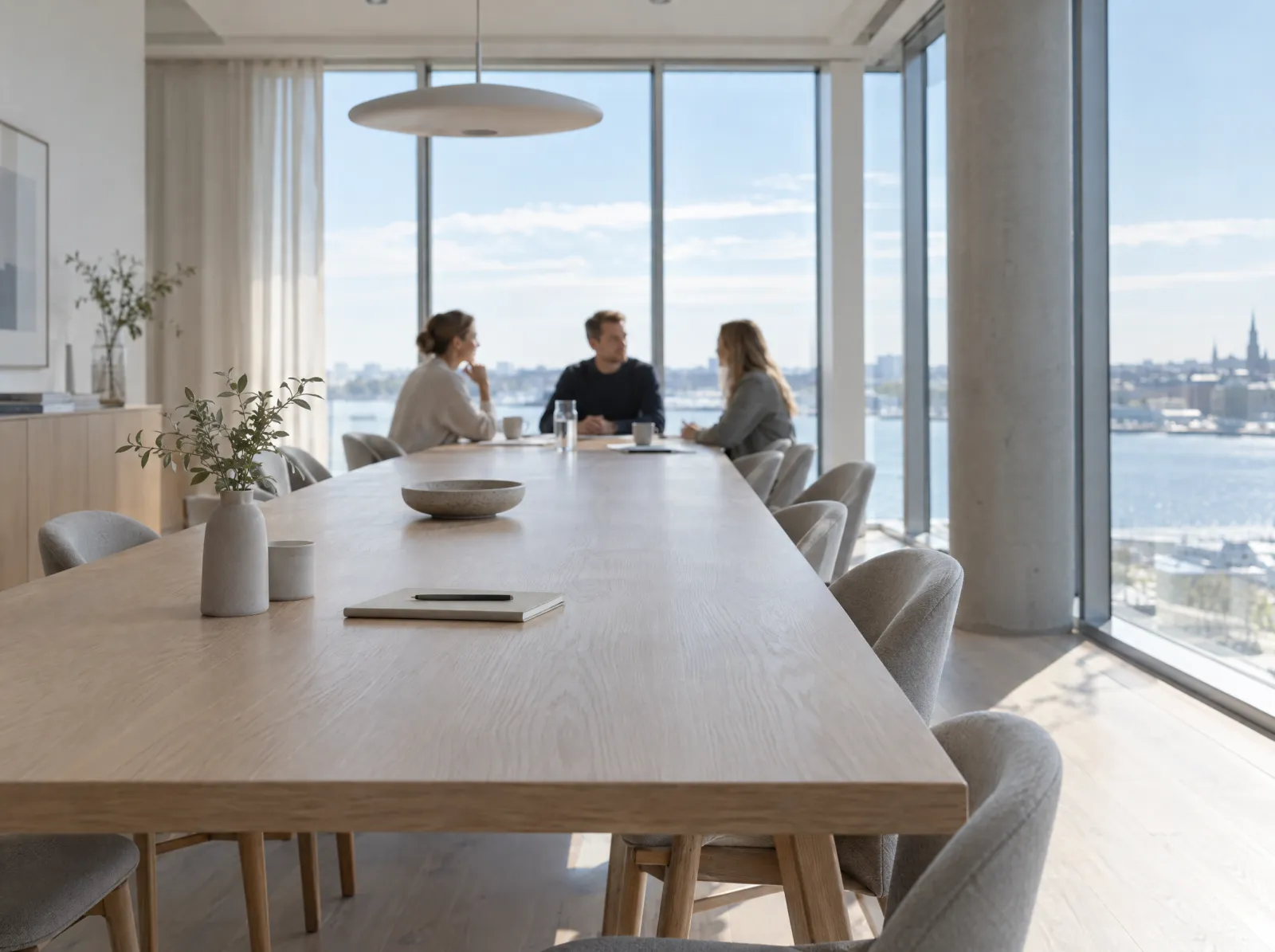 A bright meeting room with a long oak table and three colleagues talking by the window